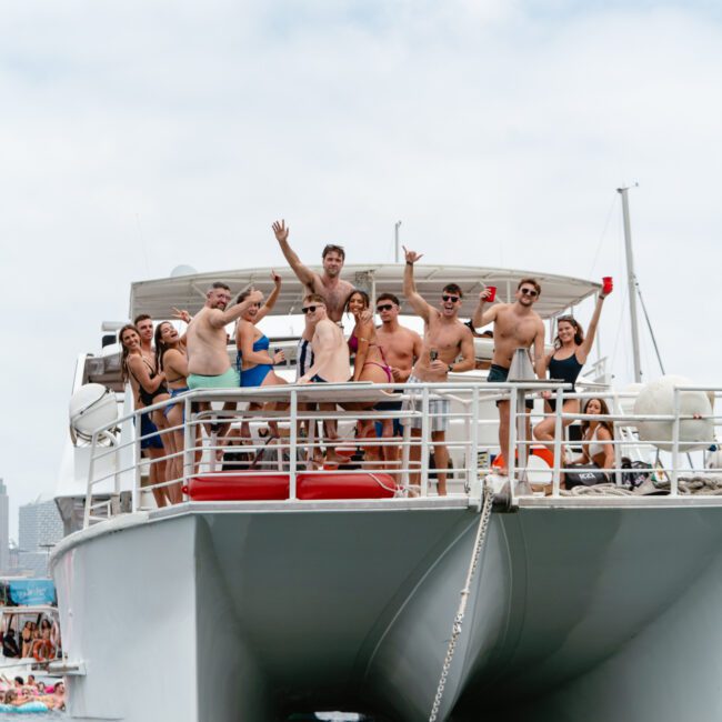 A group of people in swimwear stand and sit on the deck of a large white boat, smiling and waving at the camera. The boat, part of The Yacht Social Club Sydney Boat Hire, is anchored on a body of water with other boats visible in the background. The day looks sunny with a partly cloudy sky.