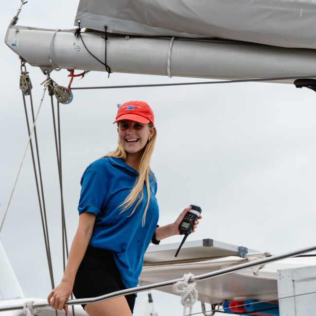 A smiling woman with long blonde hair is standing on a boat, wearing a red cap, blue T-shirt, and black shorts. She holds a walkie-talkie and seems to be enjoying her time at The Yacht Social Club Sydney Boat Hire. The background shows parts of the boat and a cloudy sky.