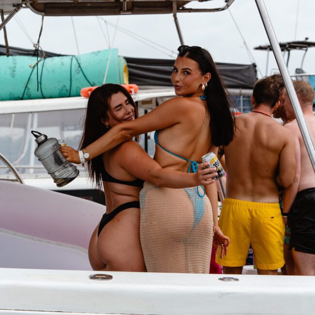 Two women in swimsuits pose on a boat from The Yacht Social Club Sydney Boat Hire. One holds a drink and smiles while the other wraps her arm around her friend, holding a can. Other people in swimsuits lounge in the background with boating equipment and another boat visible.
