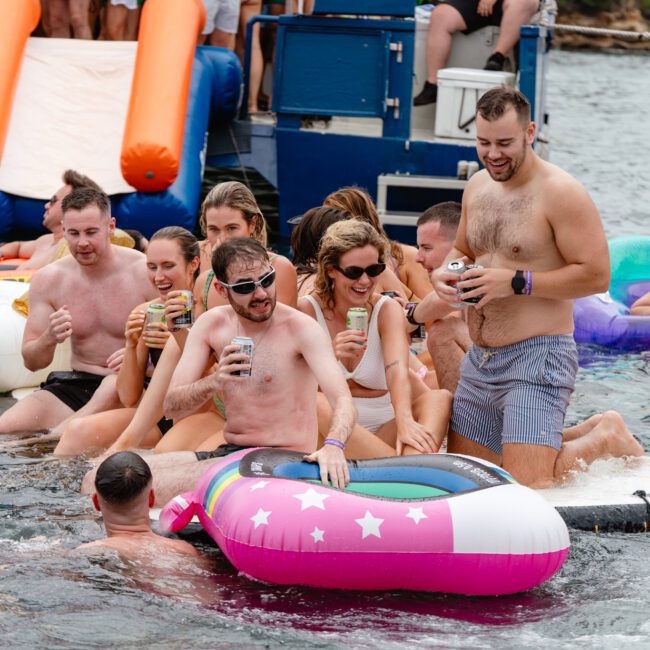 A group of people enjoy a lively time in the water, floating on various inflatable pool toys. They are smiling, drinking, and making the most of a sunny day. A boat from Sydney Harbour Boat Hire The Yacht Social Club with an orange slide is anchored behind them, enhancing the fun atmosphere.