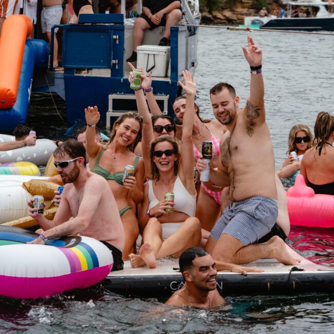 A lively group of people in swimsuits are enjoying a boat party. Several are sitting and leaning on colorful inflatable floats in the water, holding drinks and smiling at the camera. More people can be seen on the boat in the background, adding to the festive atmosphere. Welcome to The Yacht Social Club!