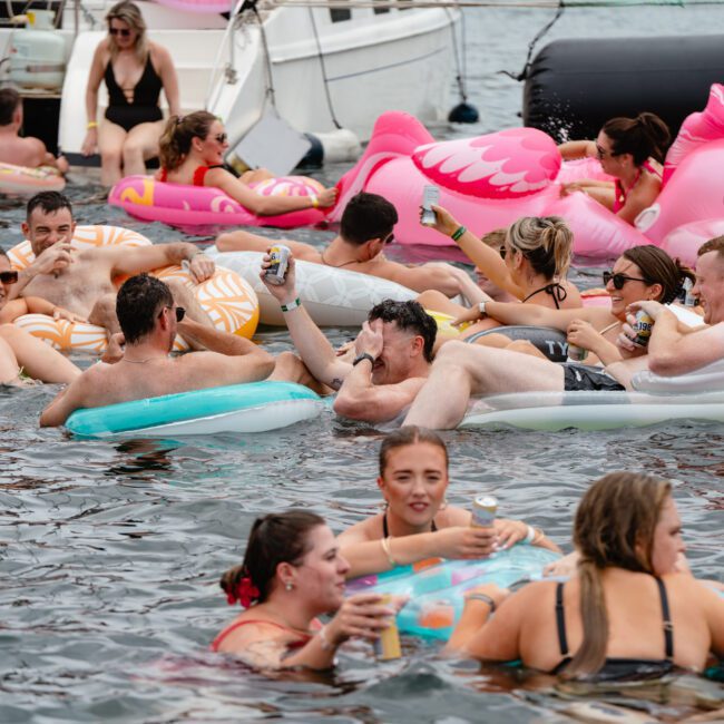 A group of people enjoying a lively party on the water with various inflatable floats, including flamingo and unicorn shapes. They are holding drinks and socializing, while boats from The Yacht Social Club Event Boat Charters are visible in the background. The atmosphere is festive and fun.