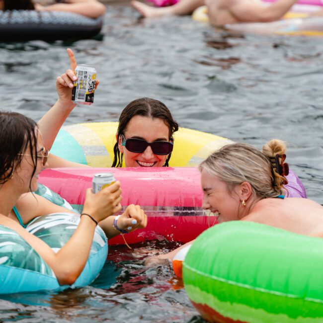 Three people enjoying a summer day in a pool, each on colorful inflatables and laughing. They hold cans of drinks, with one person raising their drink in the air. The scene is lively and fun, resembling the atmosphere of The Yacht Social Club event with other inflatables and people in the background.