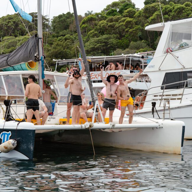 A group of people are gathered on the deck of a catamaran named "Platypus ll," enjoying the water activities. They are casually dressed in swimwear, with lush greenery in the background and another boat anchored nearby, showcasing a scene perfect for Luxury Yacht Rentals Sydney from The Yacht Social Club.