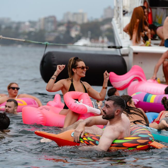 A group of people enjoying a pool party on a lake, some floating on inflatable rafts and toys, including a pink flamingo. A boat from Sydney Harbour Boat Hire The Yacht Social Club is in the background with additional people lounging. The atmosphere is lively and festive.