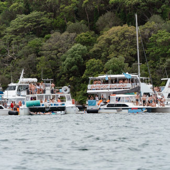Several boats, including the "Barefoot Explorer," are anchored close together on a calm body of water. People are gathered on the boats and swimming nearby, enjoying the event hosted by The Yacht Social Club. Lush green trees cover the hillside in the background.