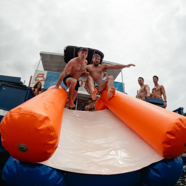 Two men are sliding down an inflatable orange and white water slide into a body of water, surrounded by cheering onlookers. Despite the cloudy weather, everyone is enjoying a fun outdoor event on the water organized by The Yacht Social Club Sydney Boat Hire.