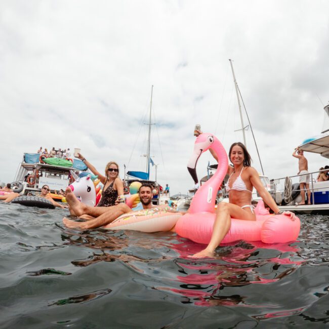 Three people sit on inflatable floats, including a unicorn and a flamingo, in a body of water during a bright overcast day. Other boats and people from The Yacht Social Club are seen in the background, creating a lively and festive atmosphere amidst the Sydney Harbour Boat Hire.