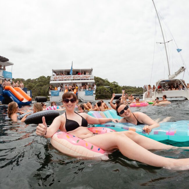A group of people are enjoying a sunny day on a body of water, lounging on inflatable floaties and boats. Two women in the foreground are smiling, giving a thumbs-up, and holding drinks. Other people are swimming and socializing around a large boat in the background at The Yacht Social Club Sydney Boat Hire event.