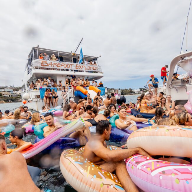 A lively crowd enjoying a boat party from The Yacht Social Club Sydney Boat Hire, with many floating on inflatable toys like doughnuts, a unicorn, and a flamingo in front of the double-decker boat. The atmosphere is festive, with coastal homes in the background.