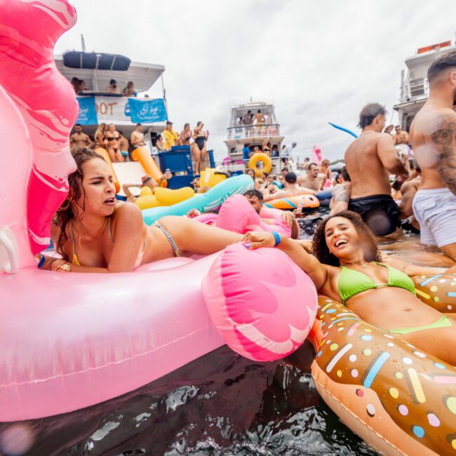 A lively scene of a pool party on the water with numerous people on colorful inflatable floats, including a pink flamingo and a doughnut. The backdrop features docked boats provided by The Yacht Social Club Sydney Boat Hire, enhancing the festive atmosphere as everyone enjoys themselves.