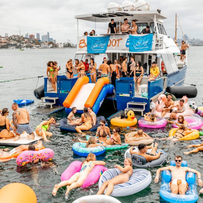 A lively scene of people partying on and around a boat on the water. The boat from The Yacht Social Club has a slide into the water, where many are floating on colorful inflatables. The sky is cloudy, and a city skyline is visible in the background.