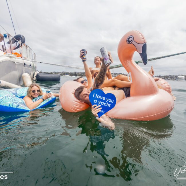 A group of people enjoying a day on the water. One person relaxes on a pink flamingo float, holding a sign that reads "I love you a yacht." Another person in a blue float and others on a boat are visible. The sky is cloudy, and the water is calm. Text: "No swipes" and "Boat Parties Sydney The Yacht Social Club".