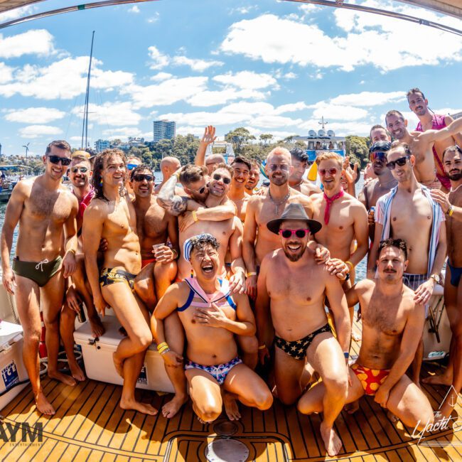 A group of people in colorful swimsuits are gathered on a Luxury Yacht Rentals Sydney boat, smiling and posing for a photo under a sunny sky. The background features other boats and a lively waterside setting. The image has a festive and joyful atmosphere.