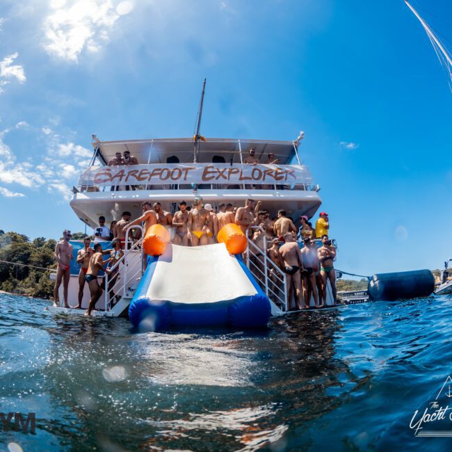 A luxury yacht labeled "Barefoot Explorer" is anchored, with people on the decks and a water slide leading into the sea. The clear blue sky and calm water create a festive and relaxed atmosphere. Sailboats and wooded shoreline are visible in the background, perfect for Yacht Social Club Event Boat Charters.