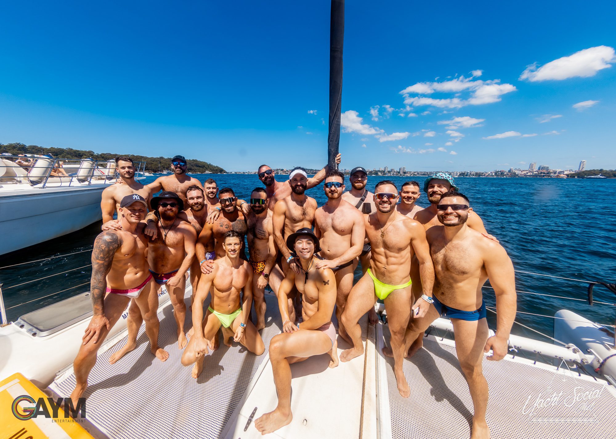A group of smiling people in swimwear poses together on a boat deck under a sunny blue sky, enjoying The Yacht Social Club Event Boat Charters. They are on the water with a scenic view of the coastline and city skyline in the background. Some are standing while others are sitting or kneeling on the deck.