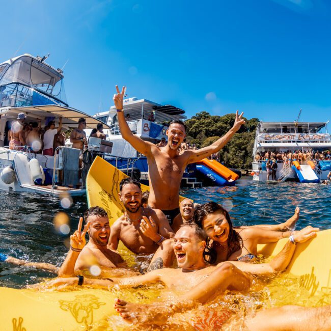 A group of people enjoy a sunny day on the water, smiling and posing on an inflatable raft. Behind them, multiple boats anchor close together, loaded with more people socializing and partying. The water is clear and blue, with a backdrop of lush green trees—an idyllic scene at The Yacht Social Club hosted by Sydney Harbour Boat Hire.