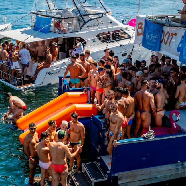 A sunny day on Sydney Harbour features a lively boat party by The Yacht Social Club. Attendees in swimsuits relax on inflatable floats and a rubber raft, while multiple boats drift on clear blue water. People mingle and enjoy the event, with city buildings visible in the background.