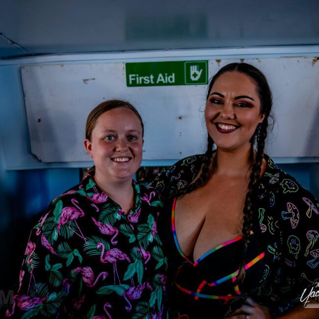 Two people smiling at the camera in a room with a "First Aid" sign behind them. The person on the left is wearing a shirt with a flamingo pattern, and the person on the right is wearing a dress with colorful symbols. The background includes "The Yacht Social Club Sydney Boat Hire.