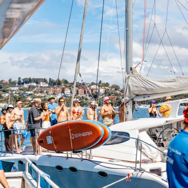 A group of people gather on a boat docked at a marina. Several individuals are shirtless and wearing sunglasses, while others are in blue shirts. A surfboard marked "SENSOR" is visible on the boat. In the background, houses and trees are seen on a hillside, highlighting The Yacht Social Club Sydney Boat Hire experience.