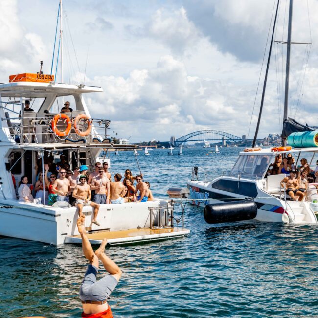 A lively scene on the water shows two yachts filled with people enjoying a sunny day. Many are in swimwear, and a person is doing a headstand dive off the back of the left yacht. In the background, a cityscape and a distinctive bridge are visible under a partly cloudy sky, highlighting The Yacht Social Club Event Boat Charters.