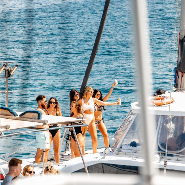A group of people in swimwear on a yacht enjoying a sunny day. Several are holding drinks and posing for photos. The sea is visible in the background, with more yachts and boats in the distance. One person takes a selfie while others smile and enjoy the moment at The Yacht Social Club Event.