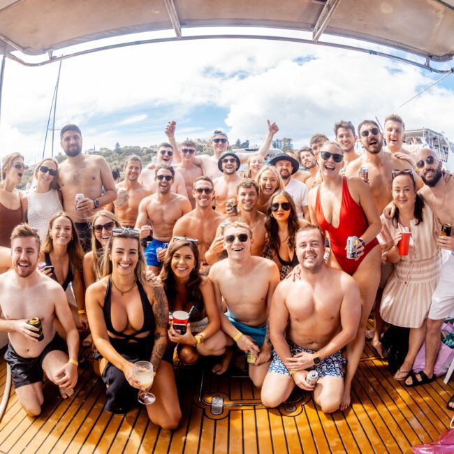 A large group of people, dressed in swimwear and summer clothing, smile and pose for a photo on a boat. The bright, sunny day features a clear blue sky and fluffy white clouds. Many are holding drinks, creating a festive and lively atmosphere at The Yacht Social Club Sydney Boat Hire event.