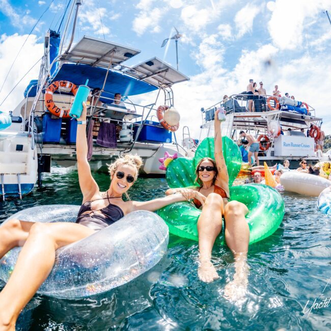 Two people float on inflatable loungers in the water, one on a clear lounger and the other on a green cactus-shaped one. They are smiling and raising their drinks. Behind them, people are gathered on boats enjoying a sunny day with The Yacht Social Club Sydney Boat Hire. The sky is blue with light clouds.