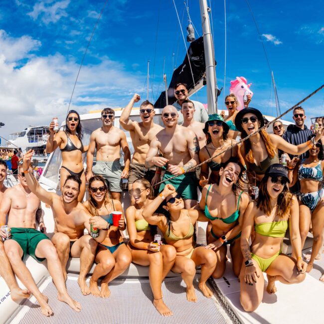 A large group of people wearing swimsuits poses joyfully on a boat deck under a sunny sky, with other boats in the background. Some hold drinks while others cheer, capturing a festive atmosphere typical of The Yacht Social Club Sydney Boat Hire events.