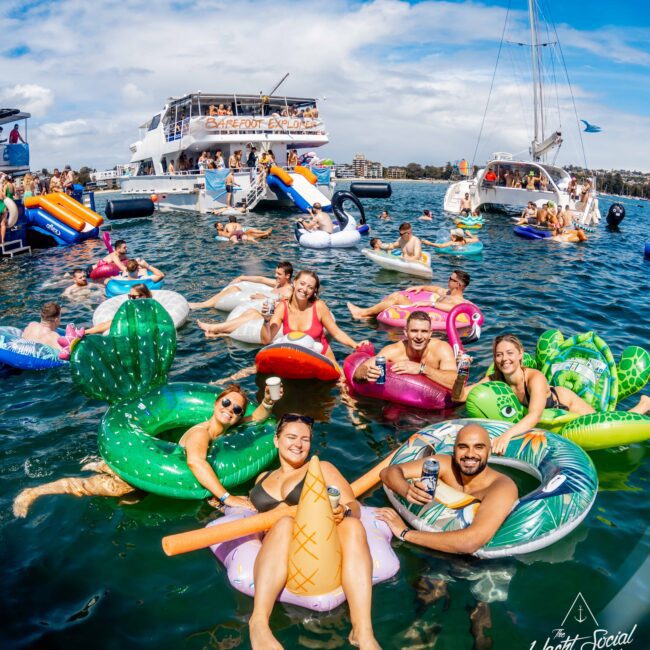 A group of people enjoying a sunny day on colorful inflatable pool floats in the water alongside yachts. They are laughing, holding drinks, and posing for the photo. One yacht in the background has a slide. The mood at The Yacht Social Club Sydney Boat Hire is festive and fun.