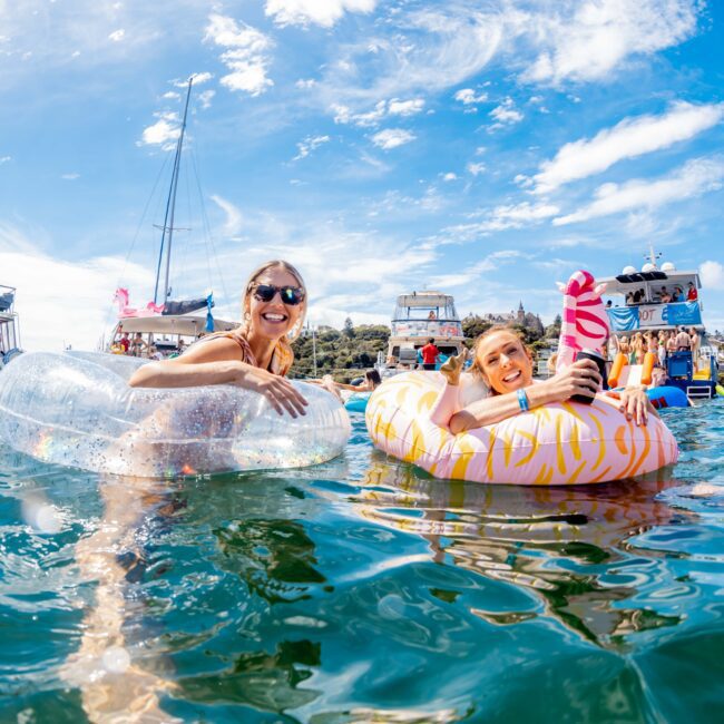 Two people floating on inflatable pool floats, enjoying the water on a sunny day. Boats filled with people from The Yacht Social Club Event Boat Charters are visible in the background, along with a vibrant blue sky. One person wears sunglasses and both appear to be smiling. The scene evokes a party atmosphere.