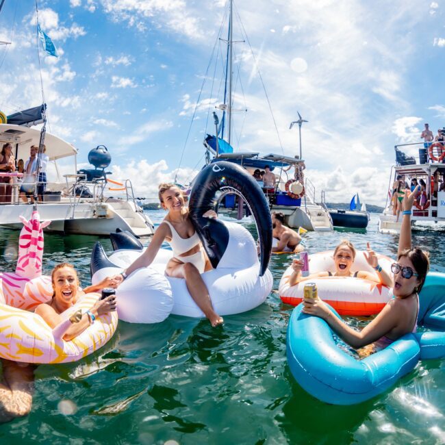 A group of people floating on inflatable pool toys, including a flamingo, swan, and raft, in a sunny and vibrant setting with boats anchored nearby in Sydney Harbour. They’re smiling, holding drinks from The Yacht Social Club, enjoying the water with a background of blue sky and clouds.