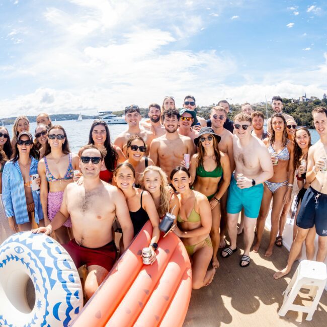 A large group of people in swimwear smiling and posing for a photo on the deck of a boat under a sunny sky. Some hold drinks, others inflatable pool items. The boat, provided by The Yacht Social Club Event Boat Charters, is on a body of water with land and buildings visible in the background.