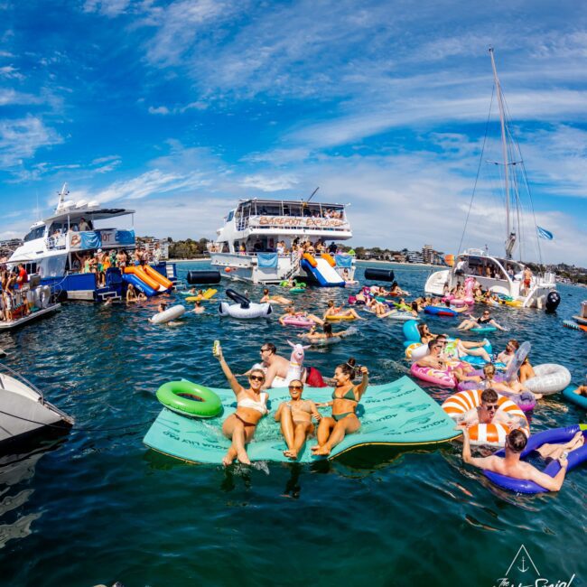 A lively scene of people at a floating party on a sunny day. The participants, some on inflatable floats, socialize and relax in the water between anchored yachts and boats. Blue skies with a few clouds are visible above. Hosted by The Yacht Social Club Event Boat Charters, the water is calm and inviting.
