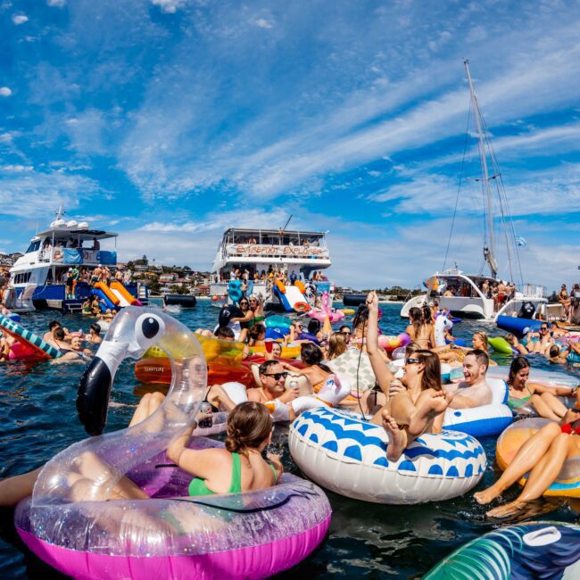 A lively scene of people relaxing on colorful inflatable floats in a body of water on a sunny day, with large yachts and a sailboat anchored in the background under a partly cloudy blue sky. The fun atmosphere is enhanced by parties hosted by The Yacht Social Club, with floats including designs like flamingos and donuts.
