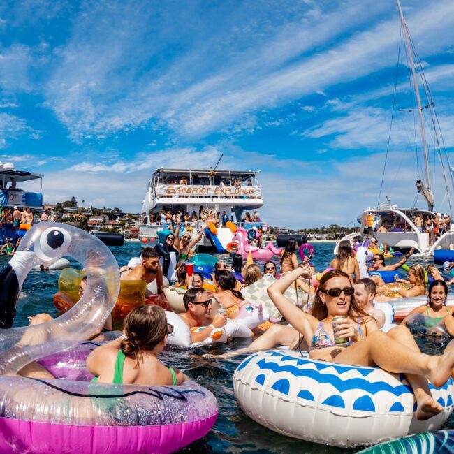 A lively scene on the water with numerous people relaxing on inflatable pool floats, including a large pink flamingo, surrounded by boats and yachts. The sky is clear with scattered clouds, and everyone appears to be enjoying a sunny day at The Yacht Social Club Event Boat Charters.