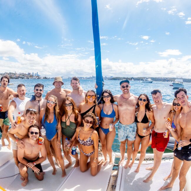 A group of people in swimwear poses together on a boat, smiling and enjoying a sunny day on the water. In the background, there are sailboats and a view of the coastline under a partly cloudy sky. Perfect for those looking into Sydney Harbour Boat Hire The Yacht Social Club events.