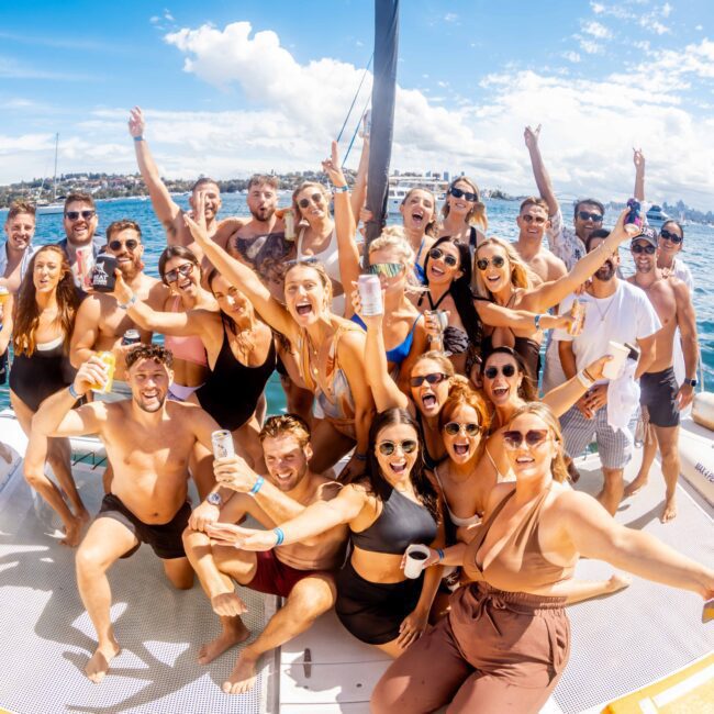 A large group of people is smiling and cheering on a boat under a bright sunny sky. They are dressed in swimwear and holding drinks, celebrating with the water and city skyline in the background. Enjoy luxury yacht rentals with The Yacht Social Club Event Boat Charters for unforgettable boat parties in Sydney.