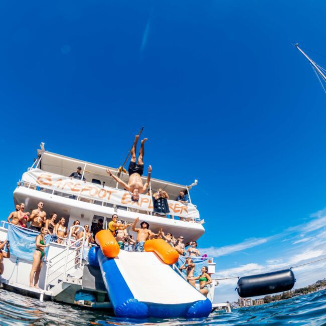 A lively group of people enjoy a sunny day on a boat named "The Spot Explorer." Some are gathered on the deck, while others slide down an inflatable water slide into the sea. The sky is clear and blue, and a nearby sailboat can be seen. It's the perfect scene for Sydney Harbour Boat Hire with The Yacht Social Club.