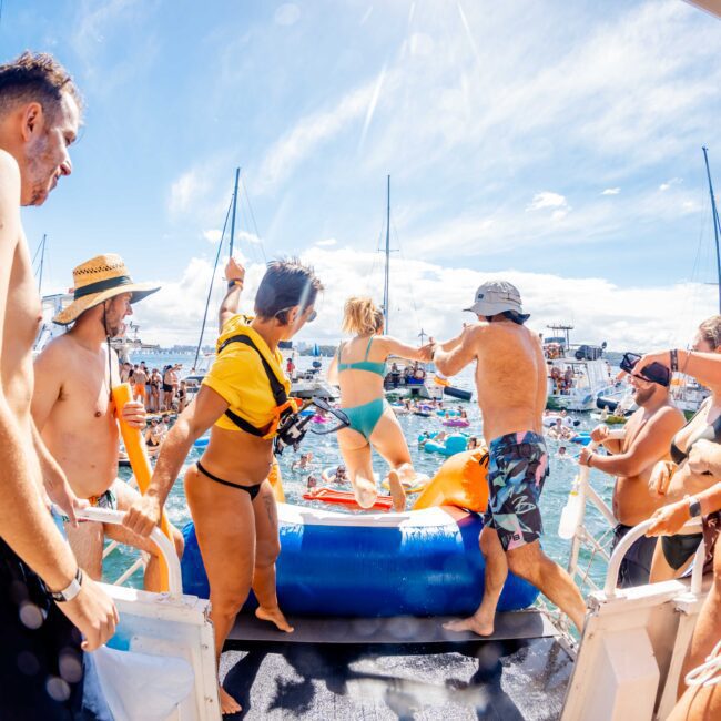 A lively scene of people on a boat enjoying a sunny day on the water. They appear to be at a party with some preparing to jump into the water where others are swimming. The background showcases clear skies and boats anchored nearby, reminiscent of events by The Yacht Social Club in Sydney Harbour.