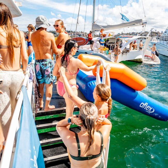 People in swimsuits and hats stand and move on a boat's ladder leading into the sea, where an inflatable float and other boats are visible. The sun shines brightly under a blue sky with scattered clouds, suggesting a lively atmosphere at The Yacht Social Club’s Sydney Harbour Boat Hire.