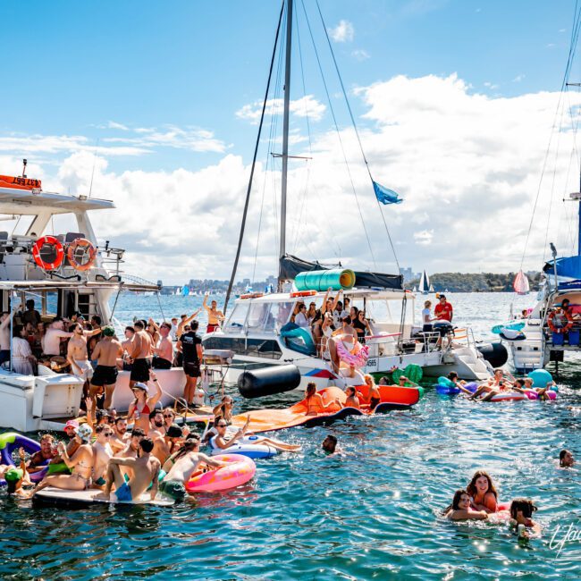 A lively scene of people enjoying a day on the water with The Yacht Social Club Sydney Boat Hire. Multiple boats are anchored close together, with individuals swimming, floating on inflatables, and socializing. The backdrop features a blue sky with scattered clouds and distant land on the horizon.