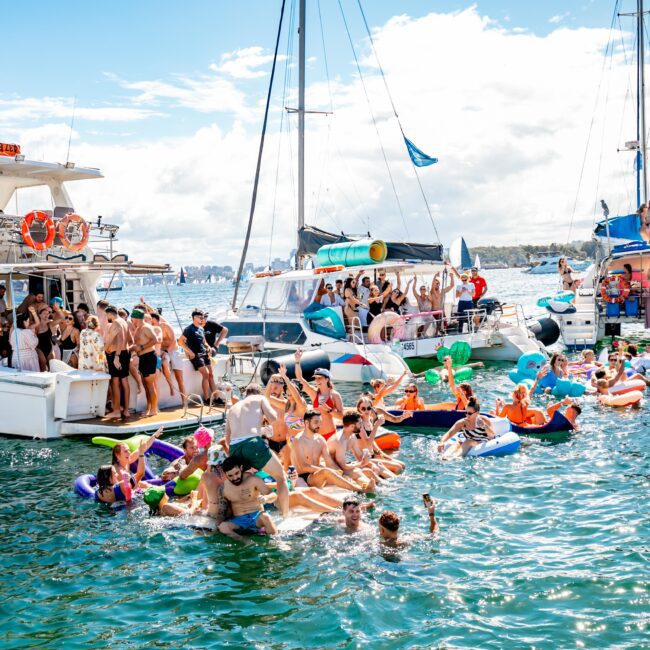 A lively gathering of people in swimsuits enjoying a sunny day on the water. Many are on inflatable floats, while others are on boats from The Yacht Social Club. The scene includes music, dancing, and general merrymaking, with multiple boats and inflatable toys adding to the festive atmosphere.