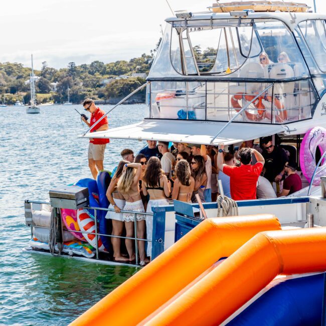 A group of people in swimsuits gather on the deck and in the water near a boat named "Sea Spirit." The Luxury Yacht Rentals Sydney boat is equipped with inflatable toys, including a water slide. The scene is festive and lively, with a backdrop of calm water and a shoreline with trees.