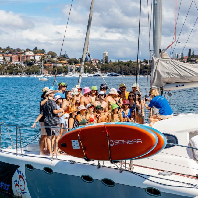 A group of people, many wearing hats and swimsuits, are gathered on the deck of a sailboat named "Barefoot". A red and white surfboard labeled "Spinera" is mounted on the boat’s railing. The boat, part of The Yacht Social Club Sydney Boat Hire, sails on a large body of water with a town visible in the background.