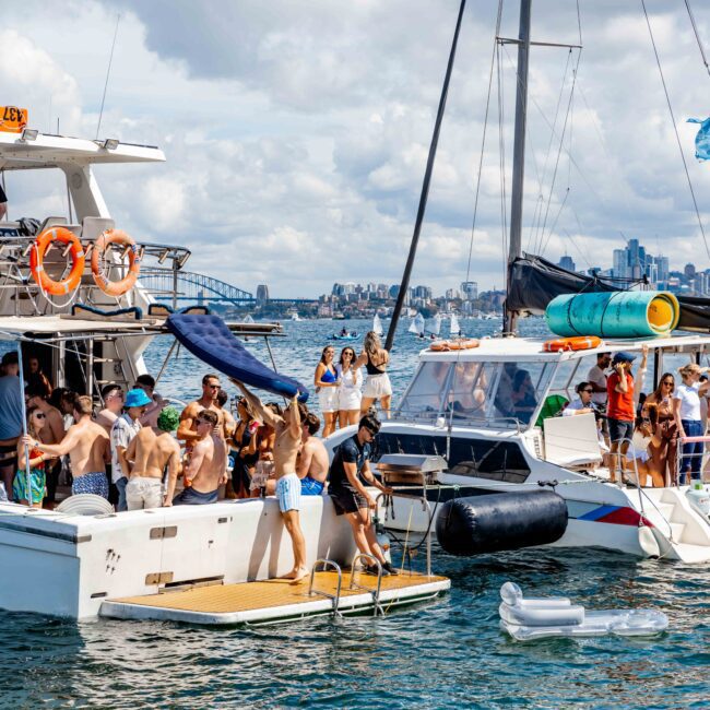 A group of people are gathered on two anchored boats and an adjacent floating platform in a sunny harbor, enjoying the vibrant atmosphere of The Yacht Social Club. Some are swimming or using inflatable floats. The background shows a city skyline with tall buildings and a partly cloudy sky.