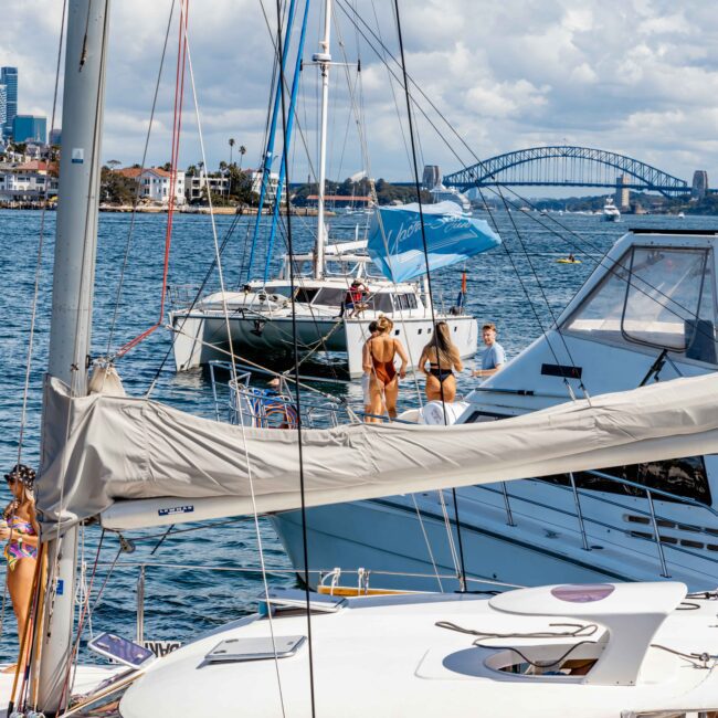 A vibrant scene of boats anchored near a city waterfront. People are enjoying the sunny day on the decks of various sailboats. In the background, a prominent bridge and urban skyline are visible under a partly cloudy sky. Some boats display colorful branding from The Yacht Social Club Event Boat Charters.