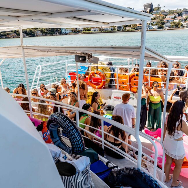 A group of people are gathered on a boat with an upper deck overlooking a body of water. The scene is lively with numerous inflatable pool floats, colorful decorations, and sunny weather. People are wearing summer attire and appear to be enjoying a Luxury Yacht Rental Sydney party hosted by The Yacht Social Club.