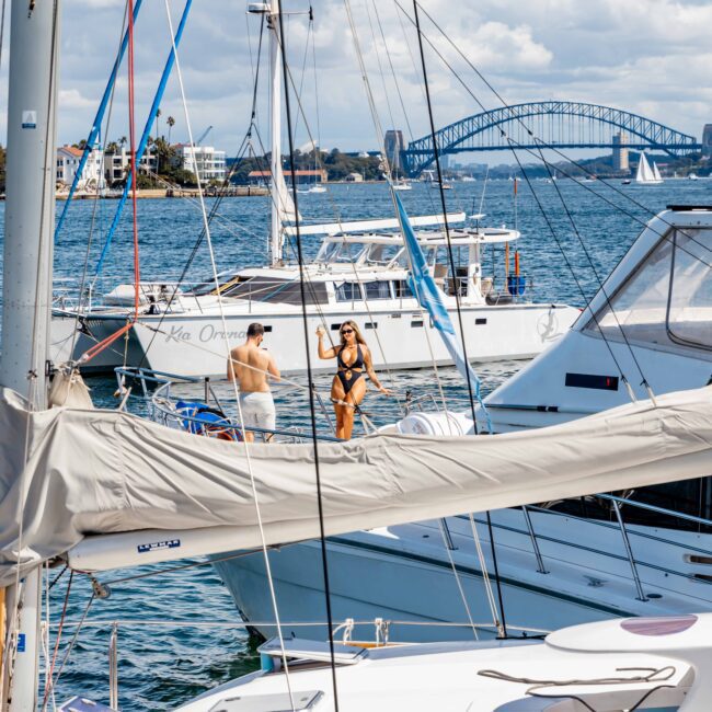 Several yachts are docked at a marina with people aboard, including a woman in a bikini sunbathing and a man standing on a nearby yacht. In the background, a bridge and several sailboats dot the partly cloudy sky. Enjoy the scene with The Yacht Social Club Event Boat Charters for an unforgettable experience.