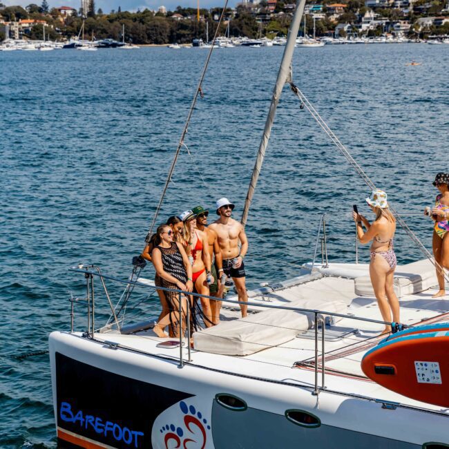 A group of people on a catamaran, some posing for a photo while one person takes the picture. The boat is on a body of water near a residential shoreline. The yacht, emblazoned with "Barefoot" on the side and featuring an orange lifeboat on deck, exudes charm similar to Luxury Yacht Rentals Sydney.